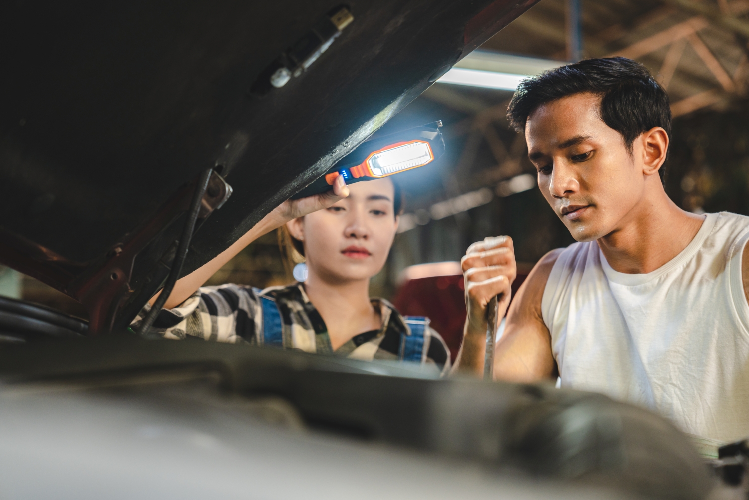 Two mechanics performing a tune up. One is holding a light so the other mechanic can see what they are doing.